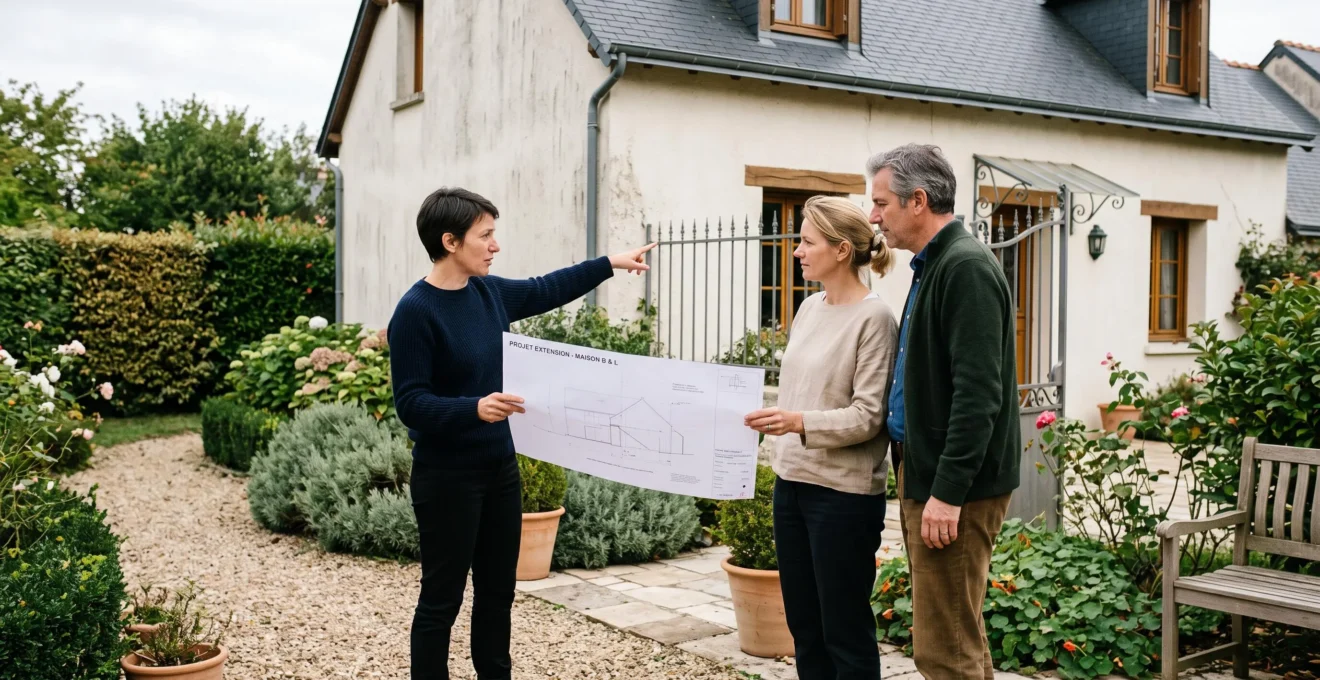 Un architecte montrant un plan d'extension à un couple de propriétaires devant leur maison, en désignant la zone concernée du projet.